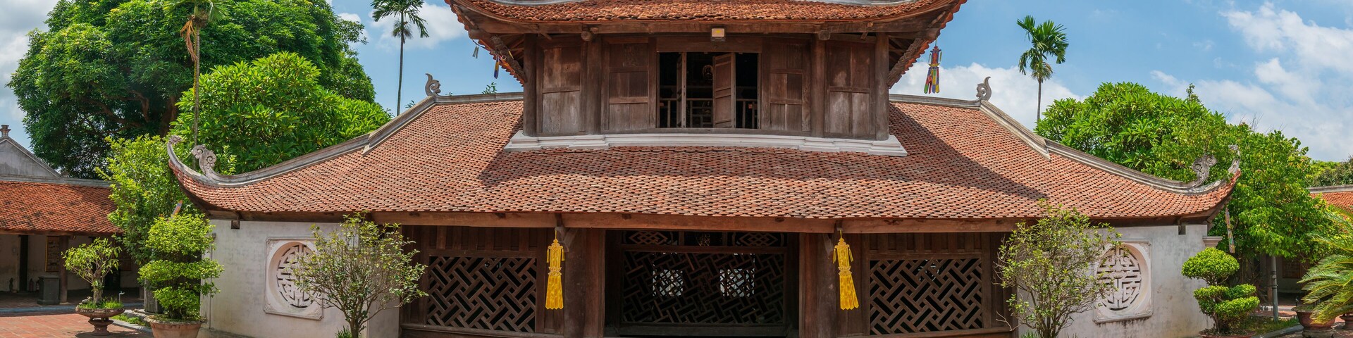 A traditional wooden building with a tiled roof and ornate decorations, surrounded by lush greenery and a paved courtyard.