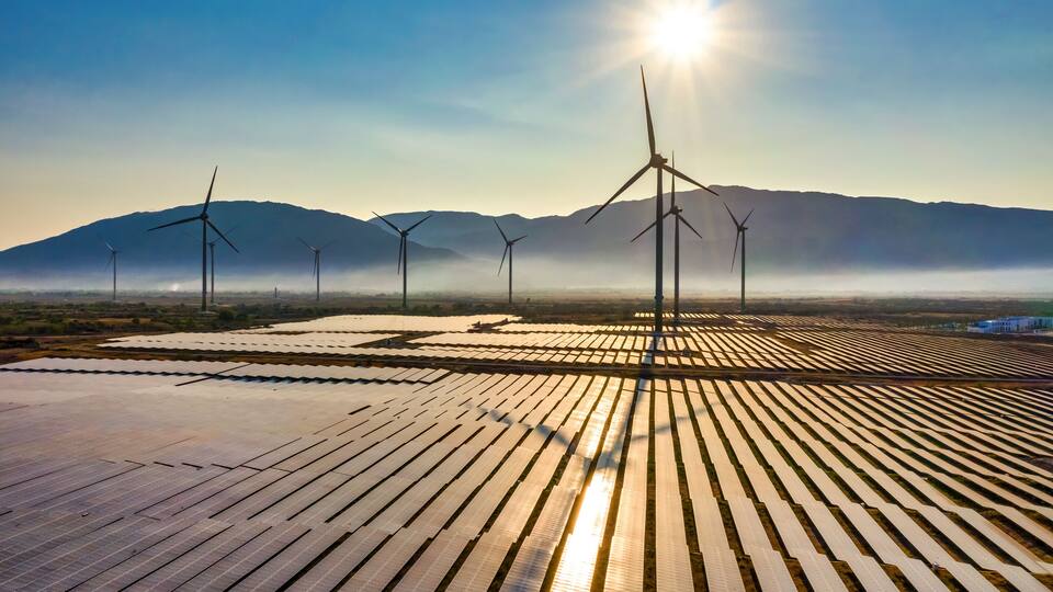 Solar panels and wind turbines in a desert landscape with mountains in the background.