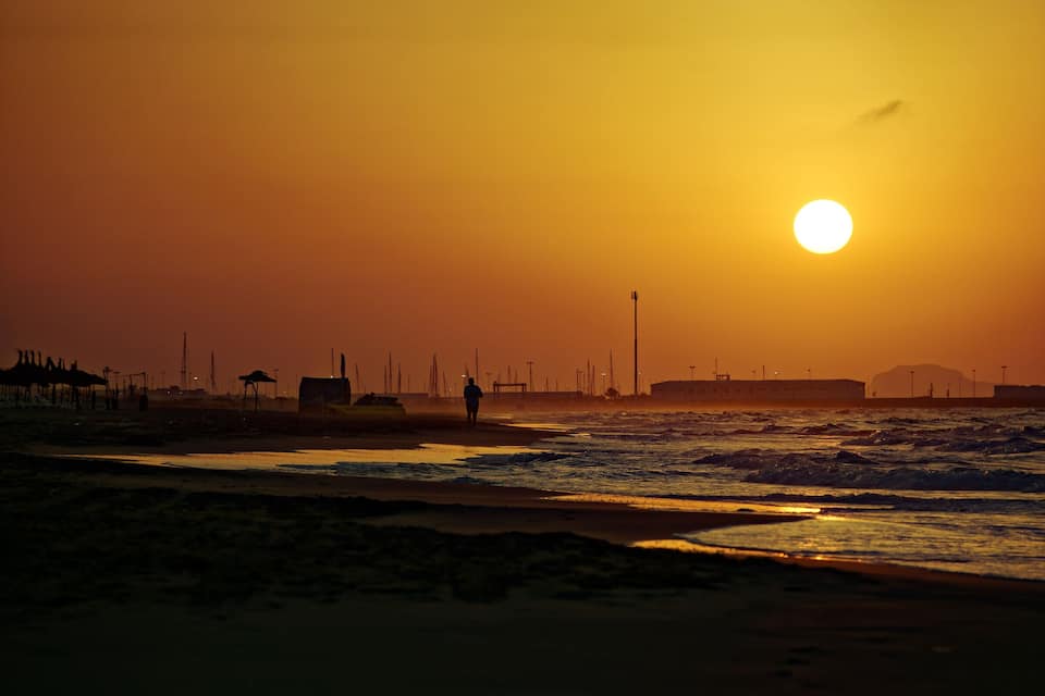 Een strand bij zonsondergang, met golven, een persoon en verre gebouwen.