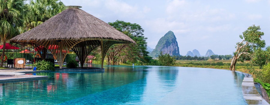 A swimming pool with a thatched-roof structure, palm trees, and distant mountains.