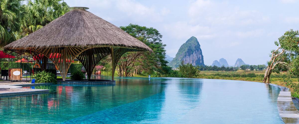 A swimming pool with a thatched-roof structure, palm trees, and distant mountains.