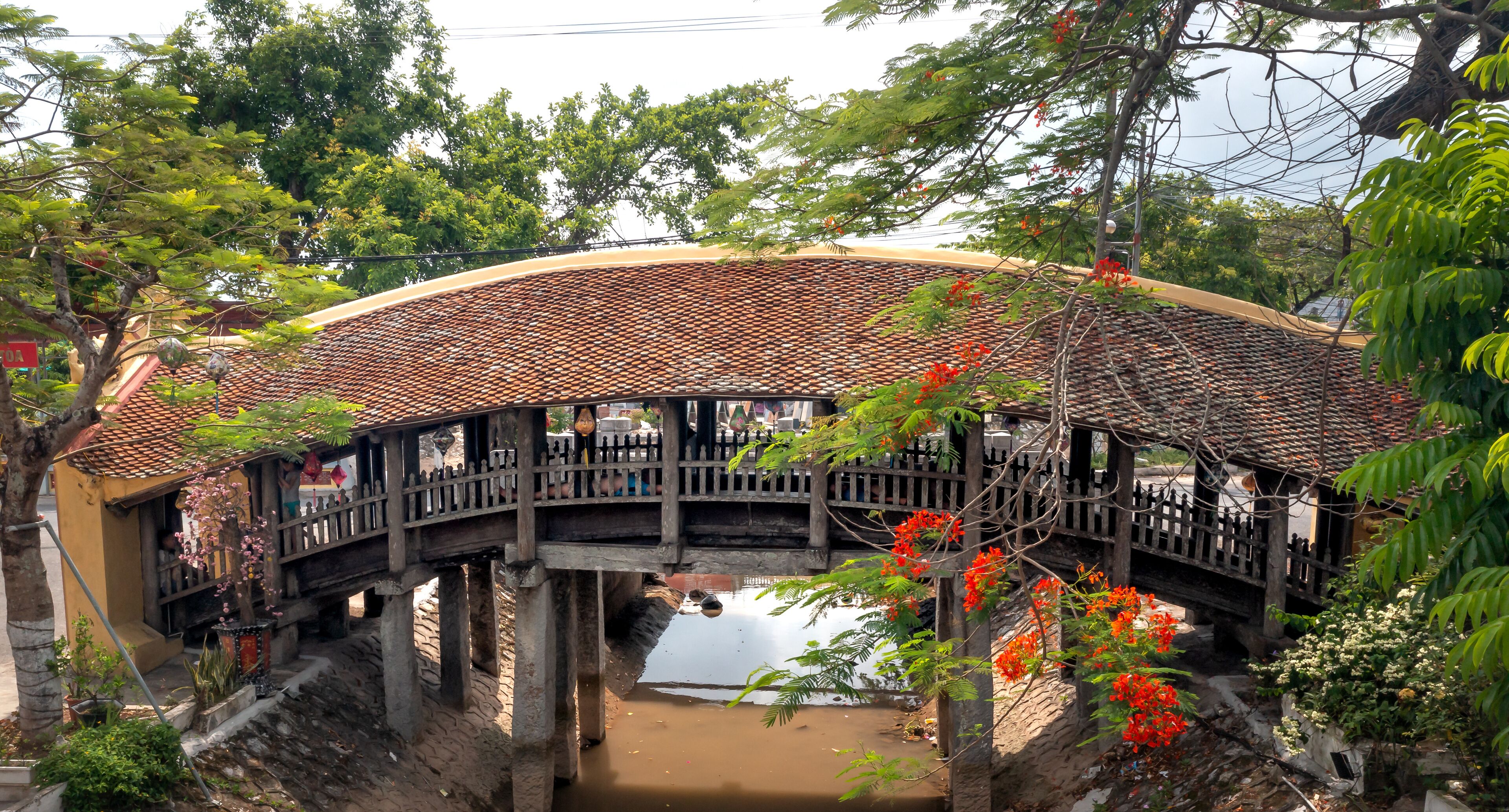 A traditional bridge with a tiled roof, supported by stone pillars, over a calm water body, surrounded by lush greenery and flowering plants.