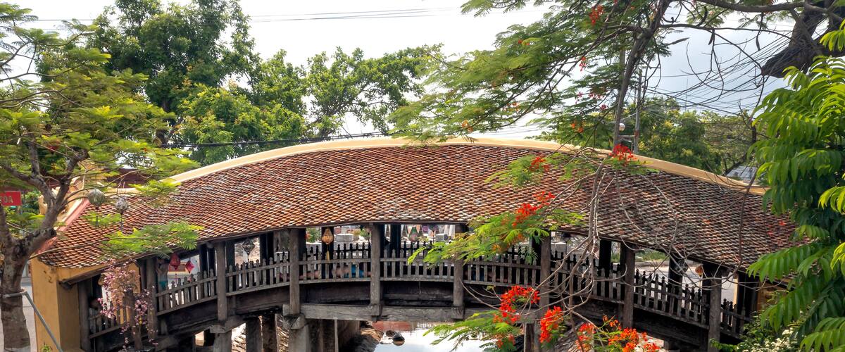 A traditional bridge with a tiled roof, supported by stone pillars, over a calm water body, surrounded by lush greenery and flowering plants.