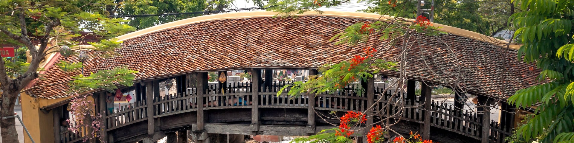 A traditional bridge with a tiled roof, supported by stone pillars, over a calm water body, surrounded by lush greenery and flowering plants.