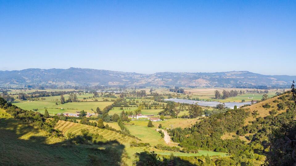 A valley with rolling hills, scattered houses, and distant mountains under a clear blue sky.