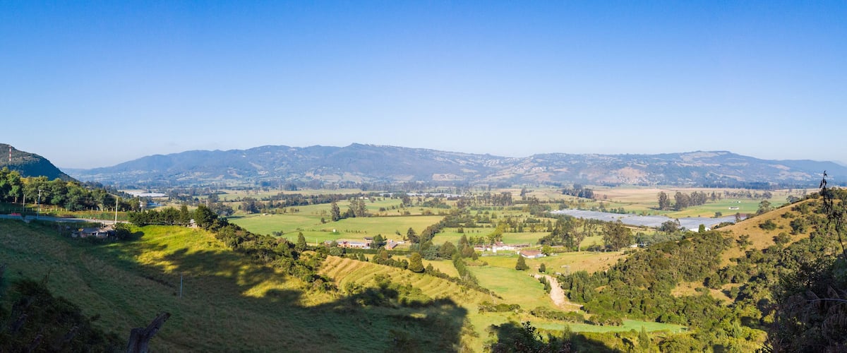 A valley with rolling hills, scattered houses, and distant mountains under a clear blue sky.