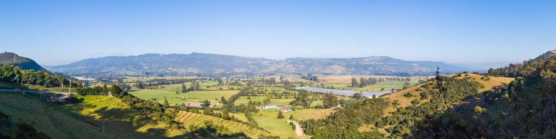 A valley with rolling hills, scattered houses, and distant mountains under a clear blue sky.