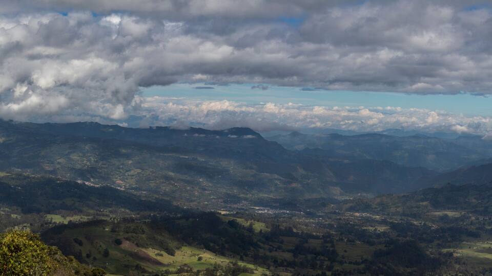 A mountainous landscape with a valley, scattered trees, and a cloudy sky.