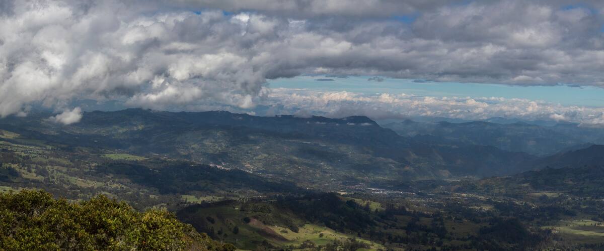 A mountainous landscape with a valley, scattered trees, and a cloudy sky.