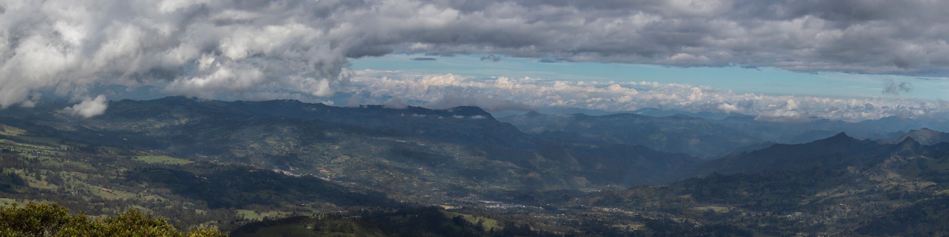 A mountainous landscape with a valley, scattered trees, and a cloudy sky.