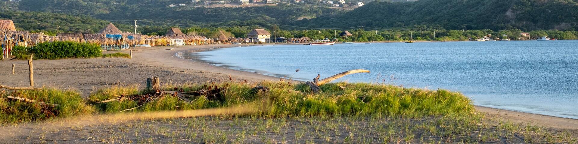 A beach with thatched-roof huts, a calm sea, and green hills in the background.