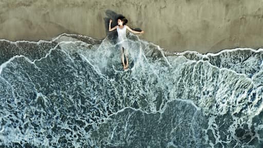 A person surfing on a wave with a sandy beach and a concrete wall in the background.