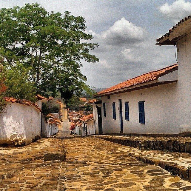 A cobblestone street lined with white buildings with red-tiled roofs, leading towards a tree-lined area.