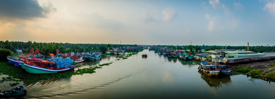 A river with numerous boats docked along the banks, surrounded by lush greenery and a cloudy sky.