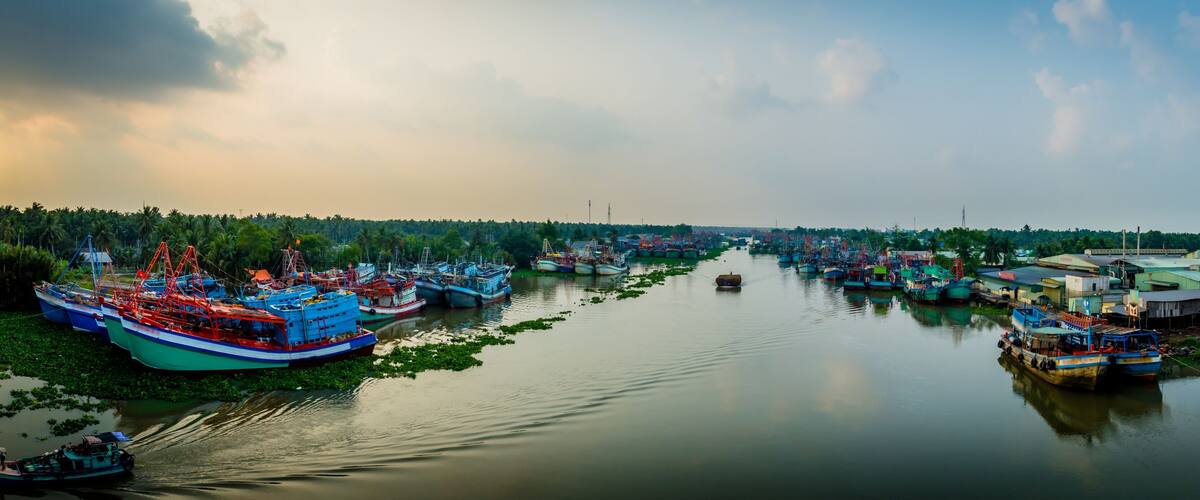 A river with numerous boats docked along the banks, surrounded by lush greenery and a cloudy sky.