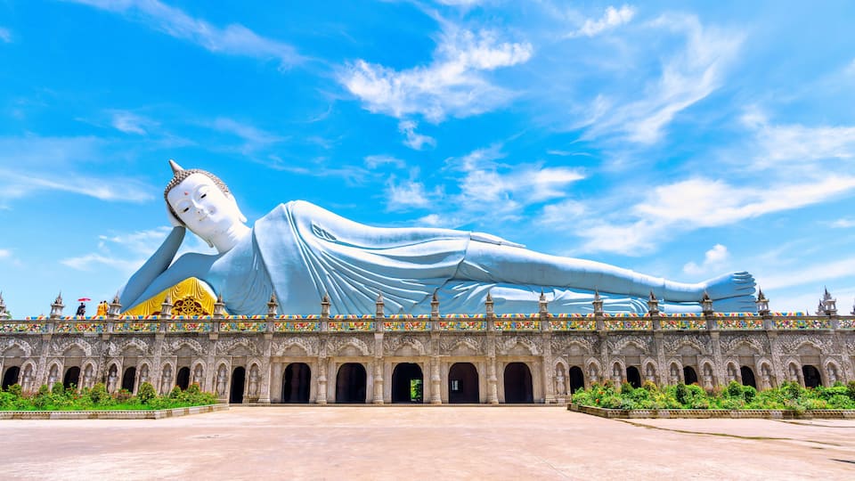 Suuri, maassa makaava Buddha-patsas temppelin edessä, ja temppelissä on koristeellinen arkkitehtuuri.