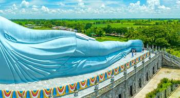 Una gran estatua reclinada de Buda con túnica azul, rodeada por una baranda ornamentada y llena de color, frente a un paisaje rural.