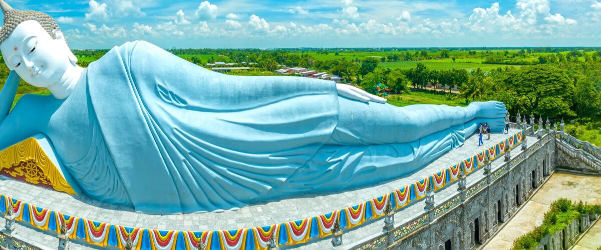 A large reclining Buddha statue with a blue robe, surrounded by a colorful, ornate railing and set against a rural landscape.