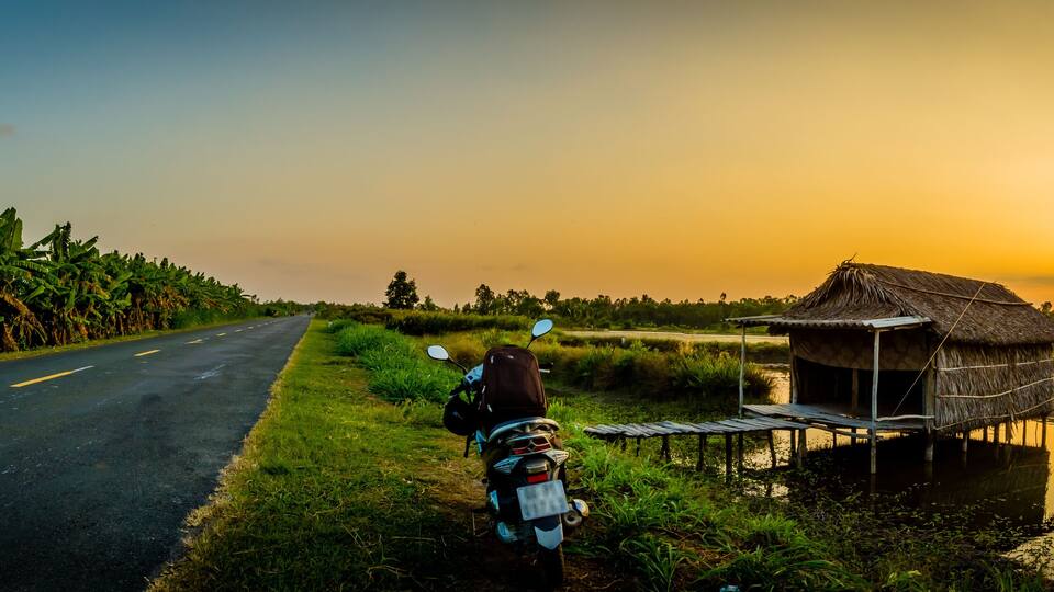 A rural road with a motorcycle parked beside a thatched-roof hut by a water body.