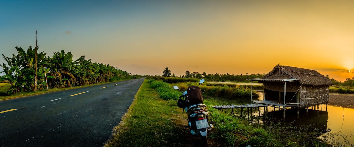 A rural road with a motorcycle parked beside a thatched-roof hut by a water body.
