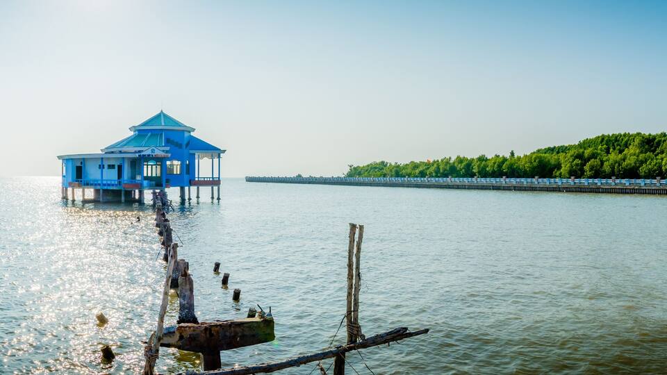 A blue pavilion on stilts in the water, a pier with a fence, and a forested hill in the background.