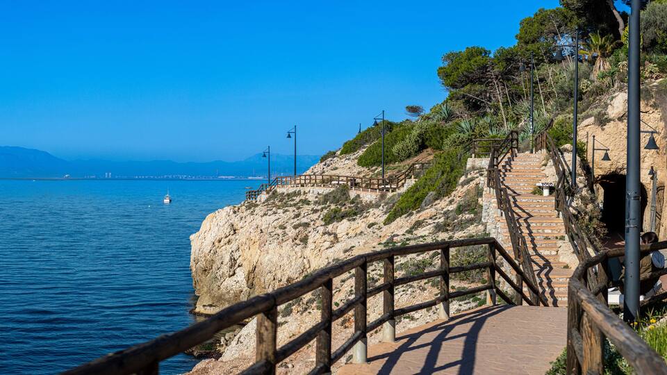 A coastal walkway with wooden railings, a stone staircase, and a clear view of the sea and distant mountains.