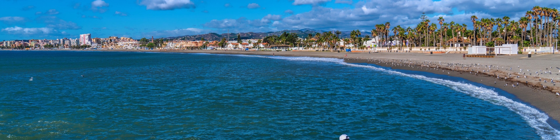 A rocky shoreline with a person taking a photo, a beach with palm trees, and a cityscape in the background.