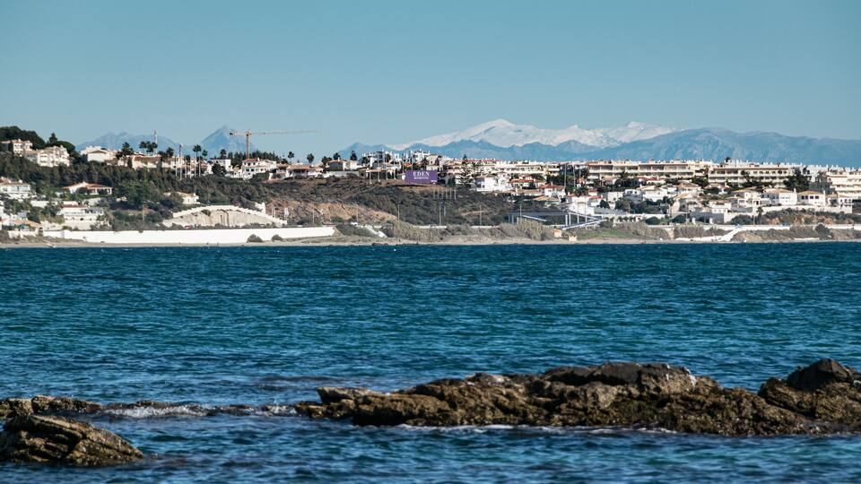 A coastal town with a clear view of the sea, rocky shoreline, and a mountain range in the background.