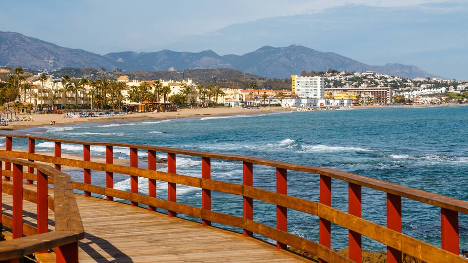 A wooden boardwalk with red railings overlooking a beach with palm trees and buildings in the background.