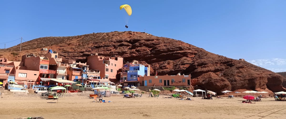 A coastal town with colorful buildings, a sandy beach, and a person paragliding in the background.