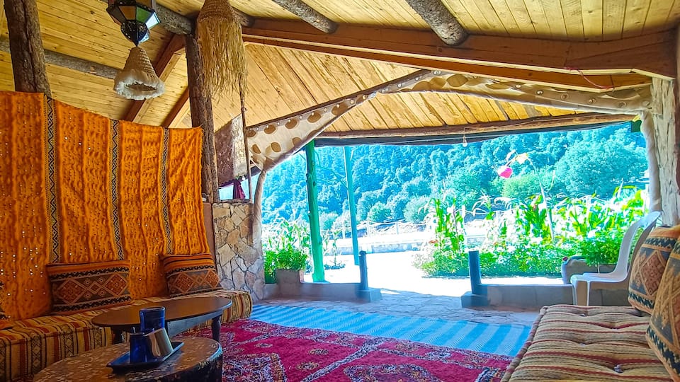 A cozy, rustic interior with wooden beams, patterned cushions, and a view of lush greenery outside.