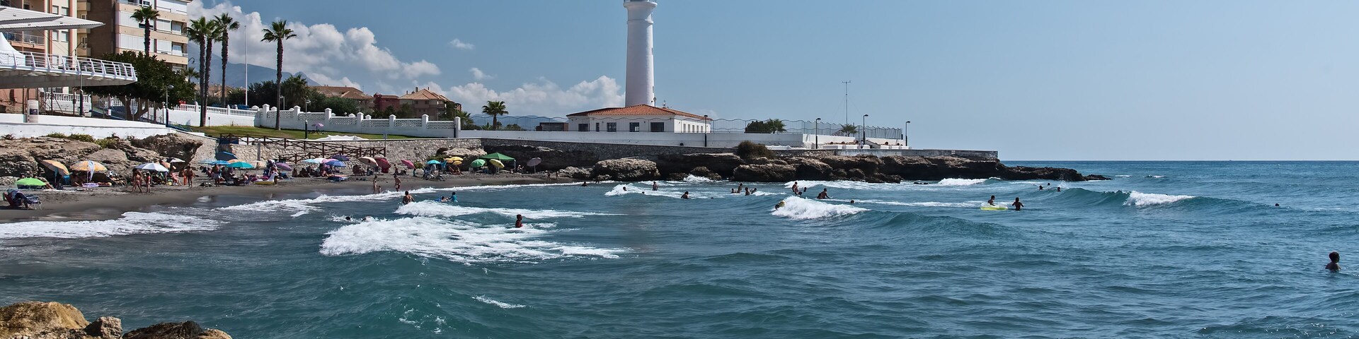Escena costera con un faro, playa, costa rocosa y edificios.