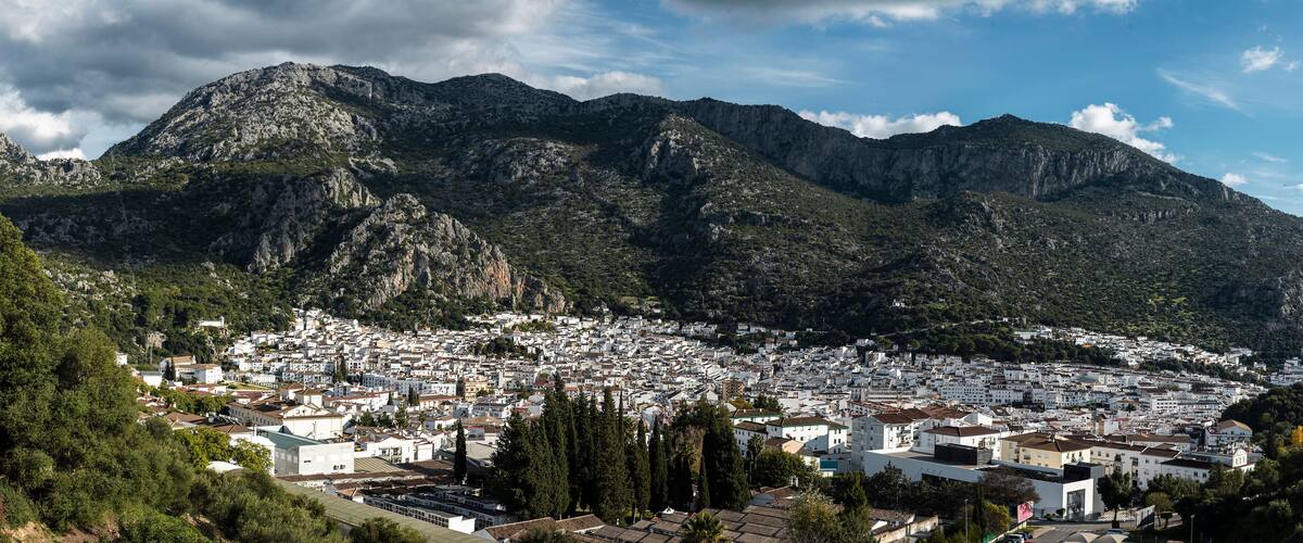 A coastal town with white buildings nestled between green hills and rocky mountains.