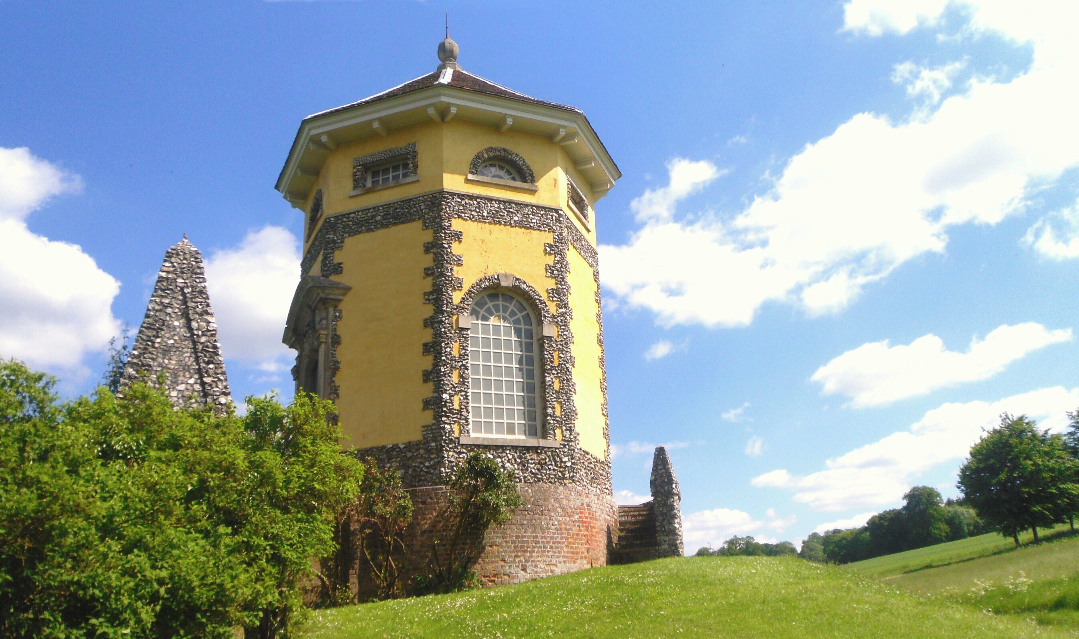A yellow tower with a stone base, surrounded by greenery and a clear blue sky.