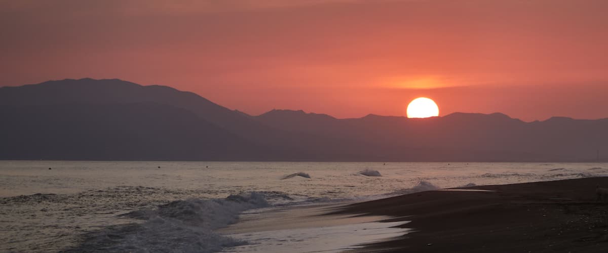 A sunset over a beach with waves and mountains in the background.