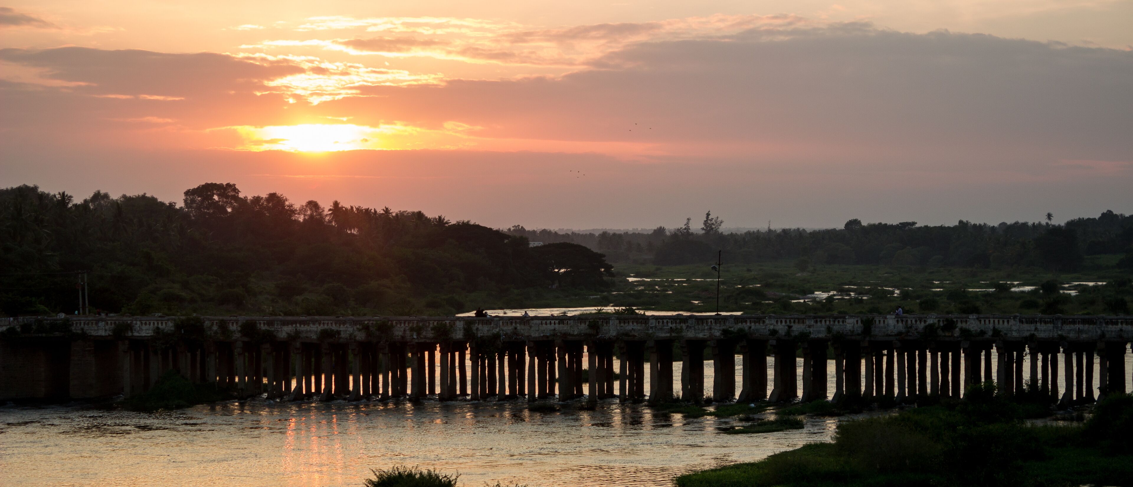 A sunset over a river with a bridge and trees in the background.