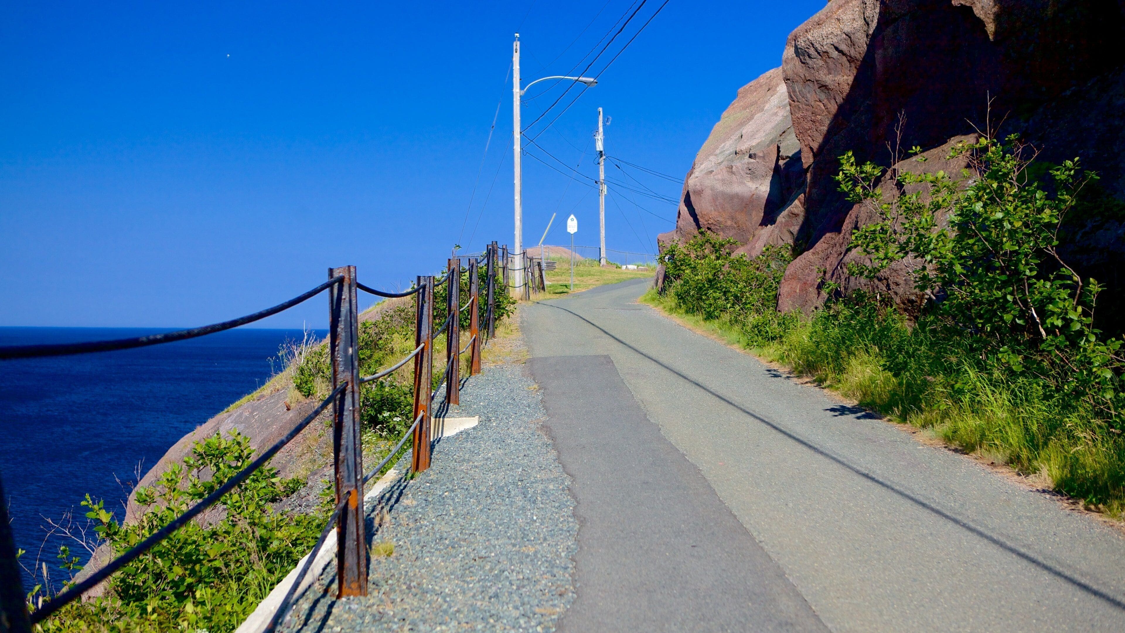 A coastal road with a guardrail, power lines, and a rocky cliff.