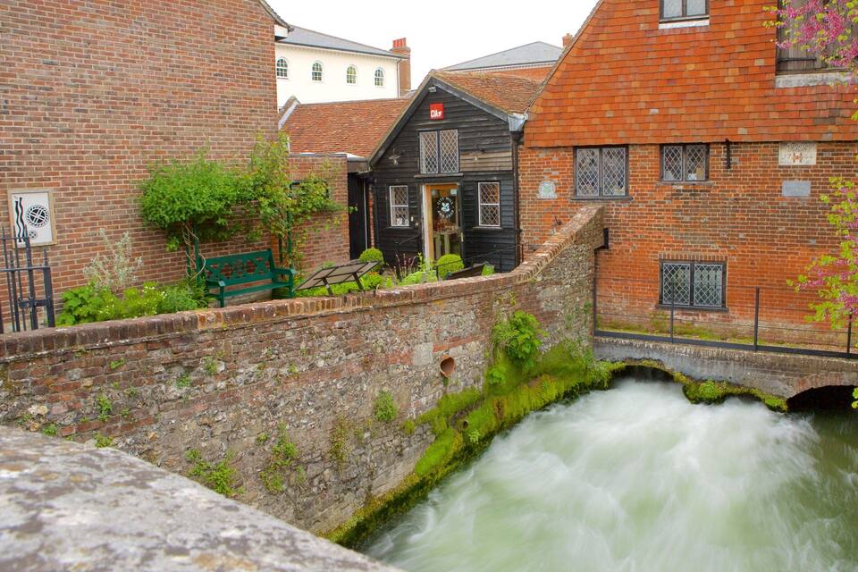 A narrow canal with a rushing water flow, surrounded by historic brick buildings and a green bench.