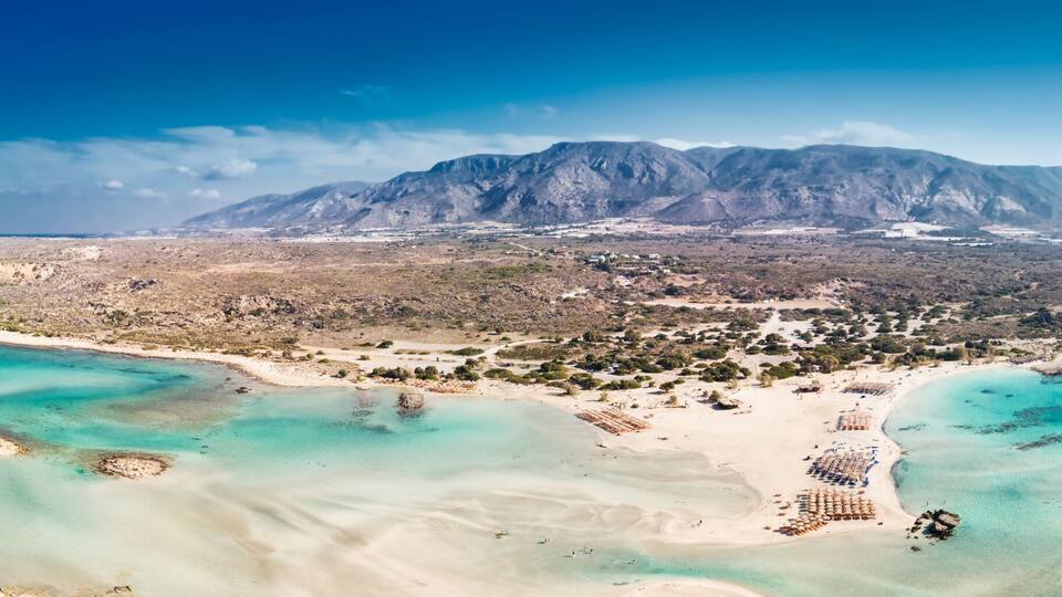 A coastal landscape with a mountain range, clear turquoise waters, and a sandy beach area.