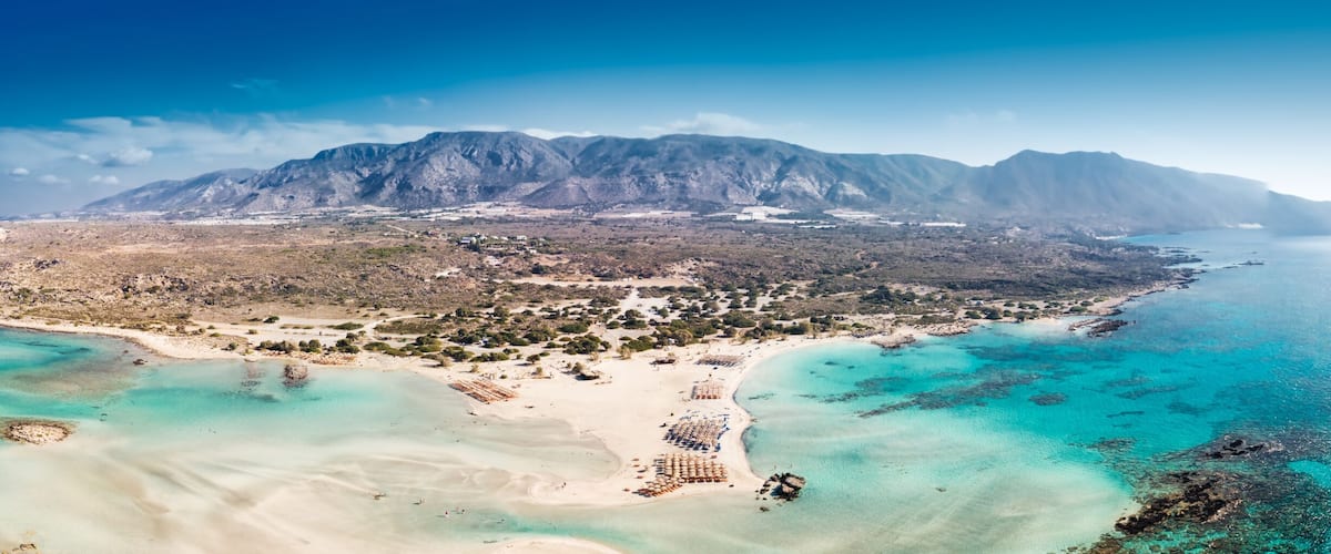 A coastal landscape with a mountain range, clear turquoise waters, and a sandy beach area.