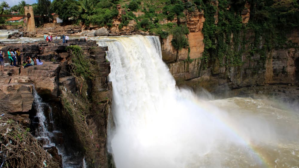 Une grande cascade, ornée d’un arc-en-ciel, entourée de falaises rocheuses et de verdure.