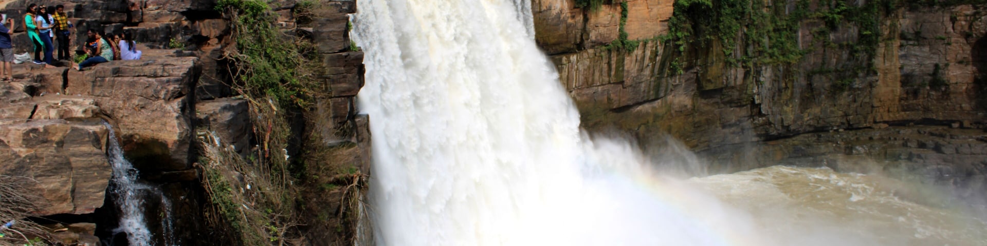 Uma grande cachoeira com um arco-íris, cercada por penhascos rochosos e vegetação.