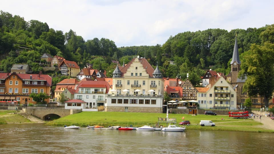 A riverside town with historic buildings, a church steeple, and boats docked along the water.