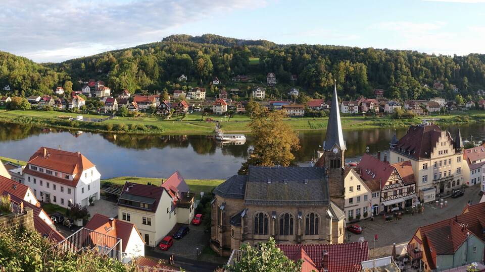 A riverside town with a prominent church, a clock tower, and a hill in the background.