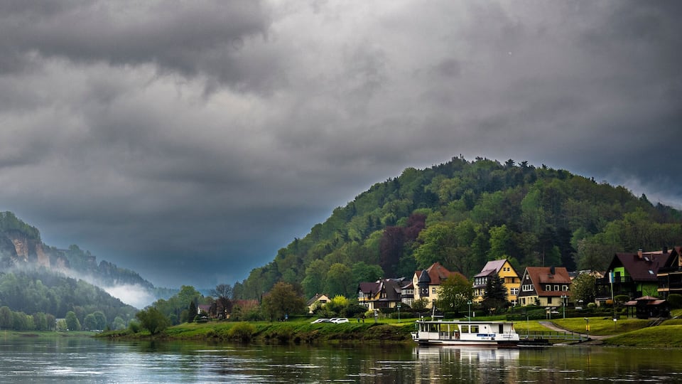 A river with houses on both sides, a boat, and a mountain with a forested peak.