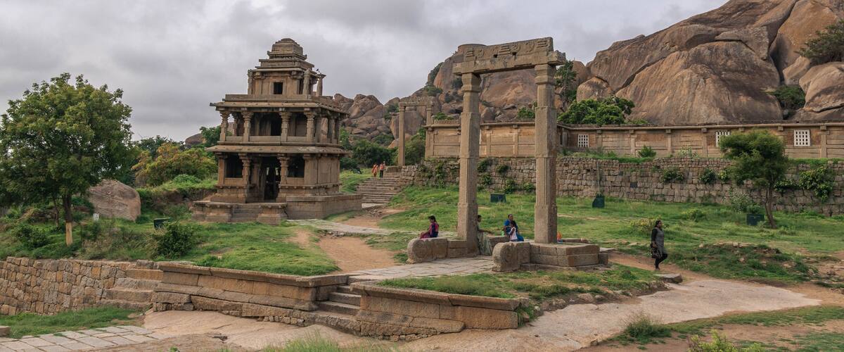 Ancient stone ruins with a temple structure and pillars, set against a backdrop of large rock formations and greenery.
