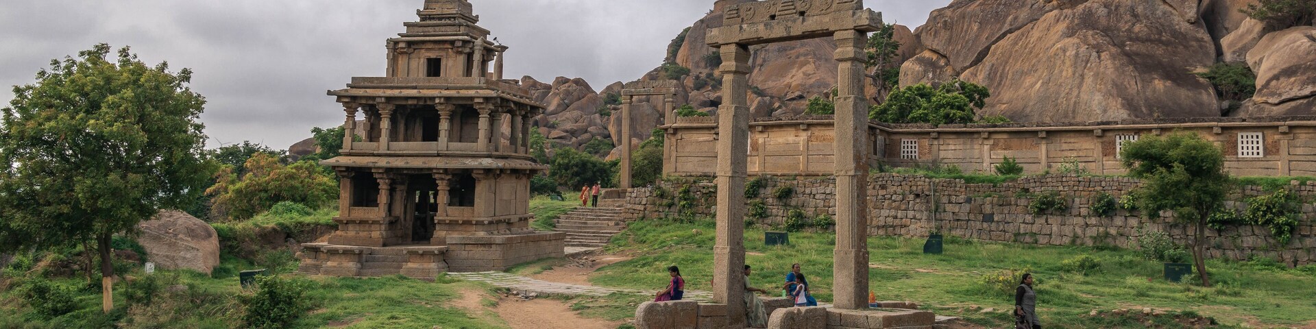 Ancient stone ruins with a temple structure and pillars, set against a backdrop of large rock formations and greenery.