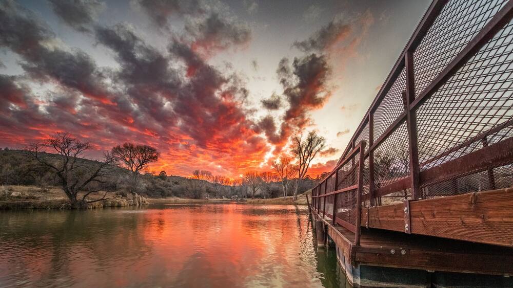 A sunset over a calm river with a wooden bridge and bare trees.