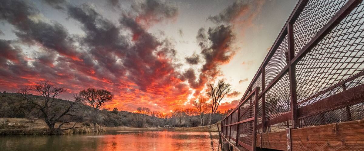 A sunset over a calm river with a wooden bridge and bare trees.
