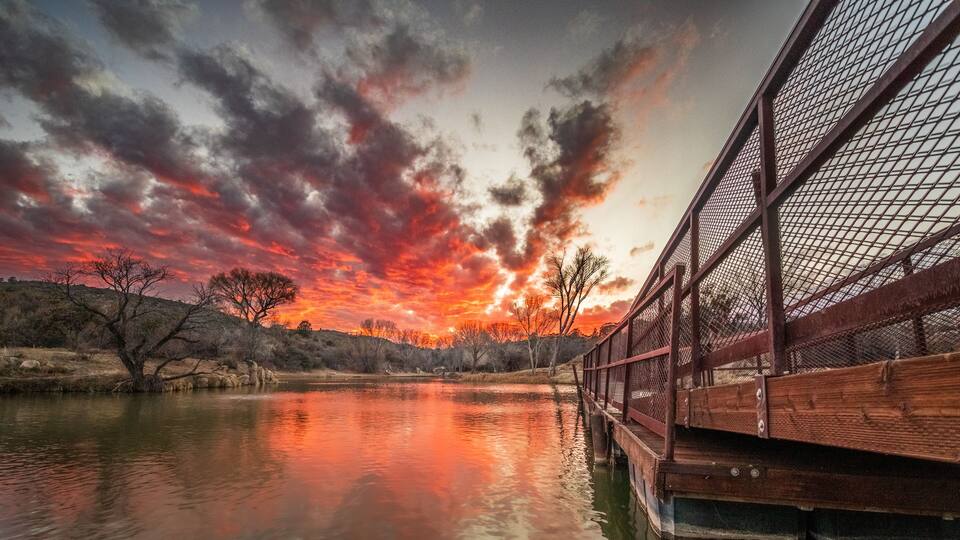 A sunset over a calm river with a wooden bridge and bare trees.